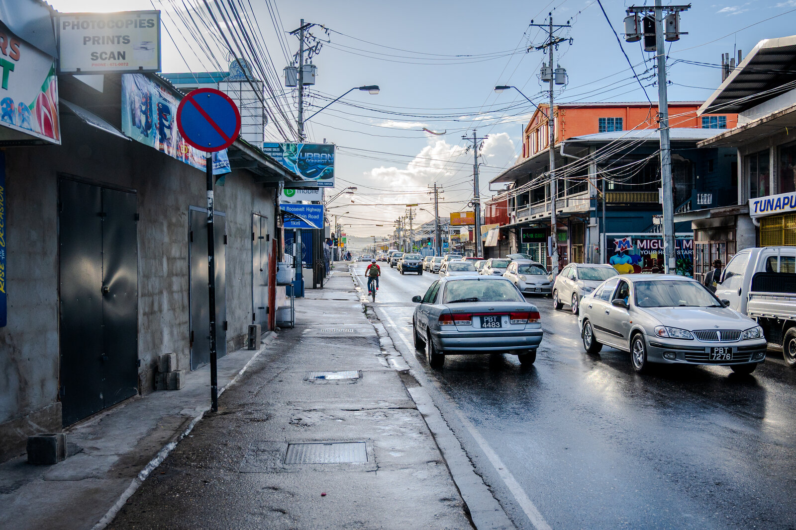 Eastern Main Road in Tunapuna on a typical afternoon, with shopfronts, taxis, and a cyclist moving down the road.